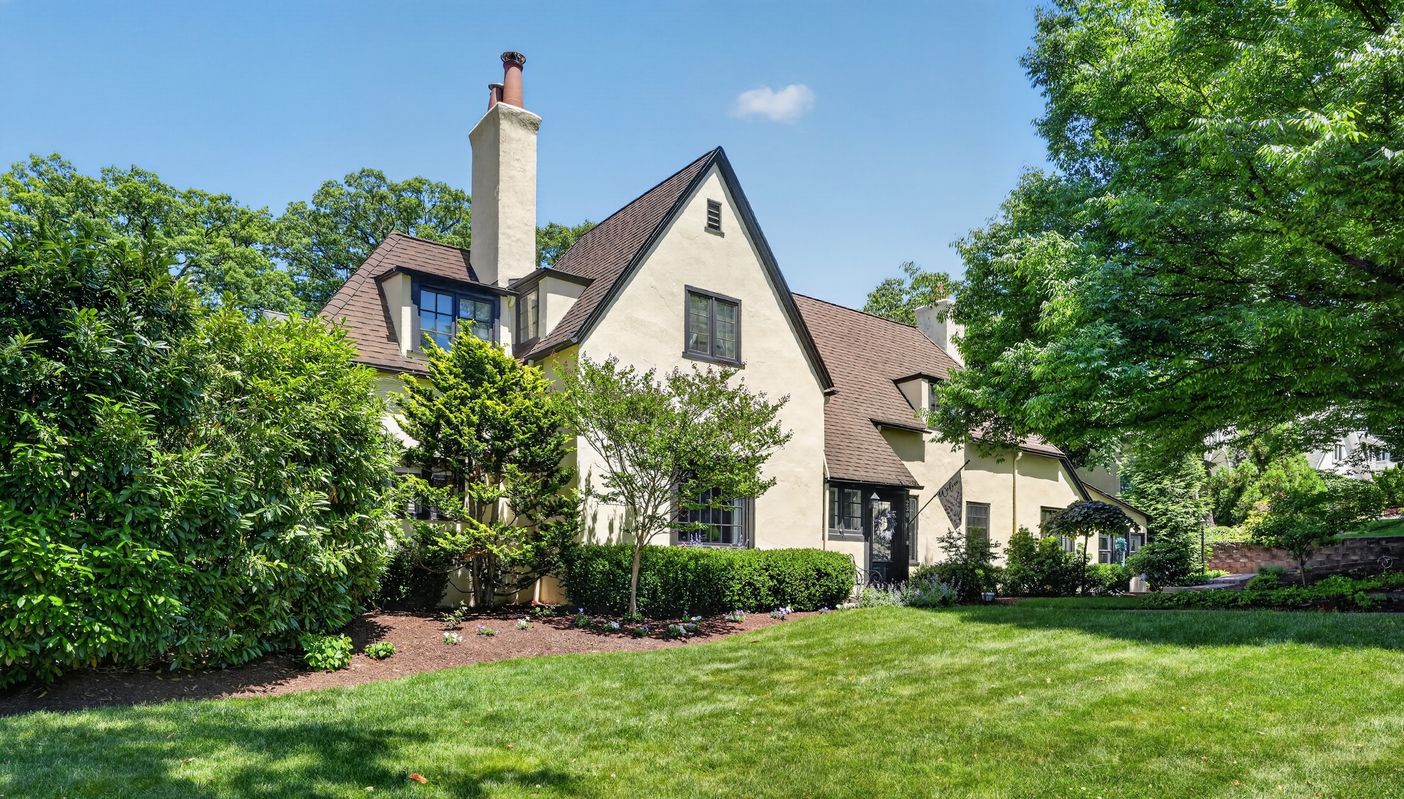 Cream-colored cottage with steep brown roof, chimneys, and lush garden under a clear blue sky.