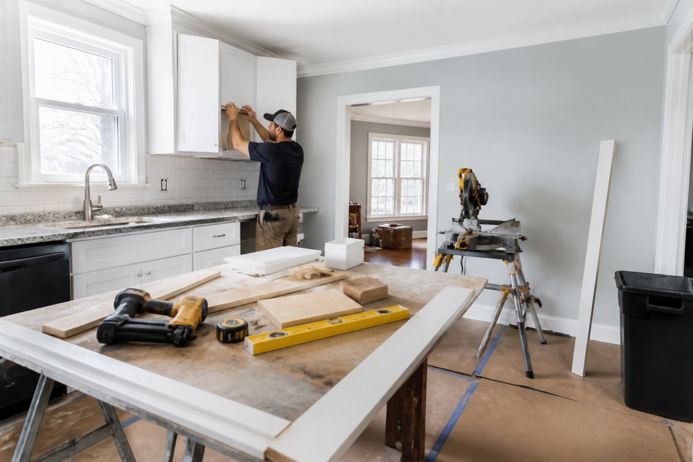 Construction worker installs white upper cabinets in a bright kitchen, granite counters and a large window nearby.