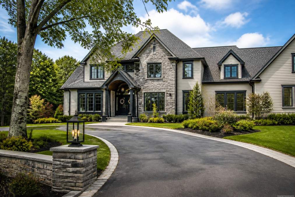 Two-story stone-and-siding house with a curved asphalt driveway and well-manicured lawn and shrubs in front.